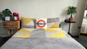 Un lit recouvert d’une couvre-lit à motifs gris et jaunes, un oreiller orné du logo du métro londonien, une cafetière rouge et une plante en pot.