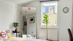 Une chambre à coucher avec un lit, une plante en pot, un radiateur, une horloge et une fenêtre donnant sur des immeubles.