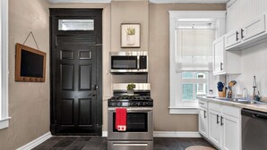 Una cocina con puerta negra, electrodomésticos de acero inoxidable, armarios blancos y una ventana con persianas.