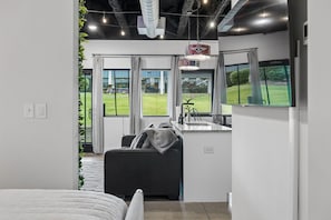Een moderne hotelkamer met een groot bed, een bank, een kitchenette en grote ramen met uitzicht op een gazon.