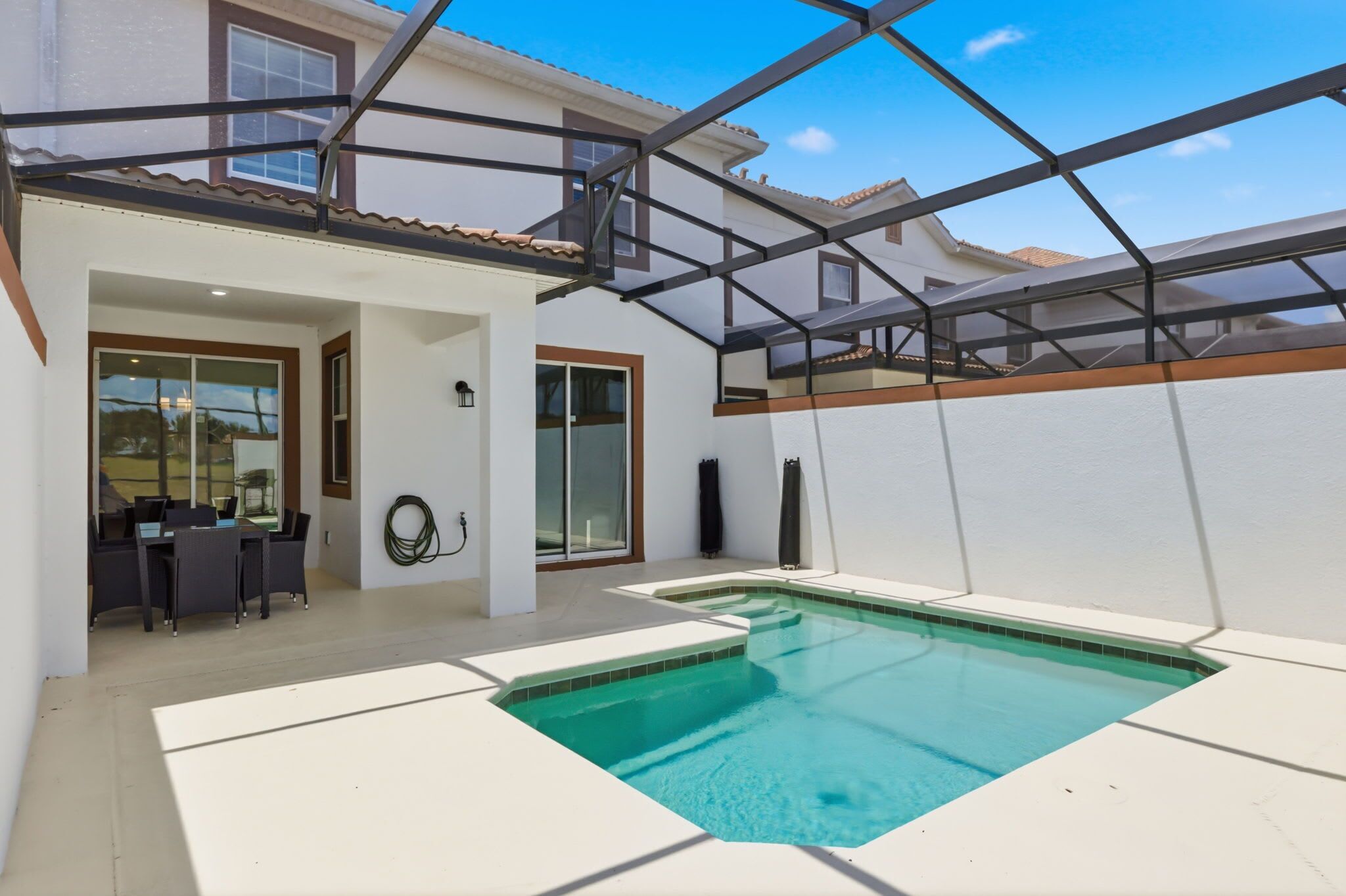 A swimming pool with a glass enclosure, a patio area with a table and chairs, and a building with a tiled roof.
