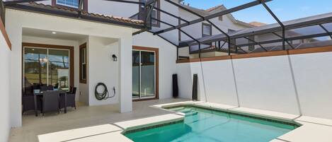A swimming pool with a glass enclosure, a patio area with a table and chairs, and a building with a tiled roof.