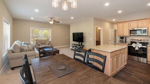 A modern kitchen with built-in appliances, a dining area with a wooden table and chairs, and a living room with a sofa and TV.