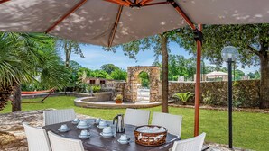 Un patio al aire libre con una mesa lista para comer, rodeado de sillas y cubierto por una sombrilla grande.