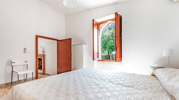A bedroom with a large bed, a window with an open shutter, a chair, and a small table.