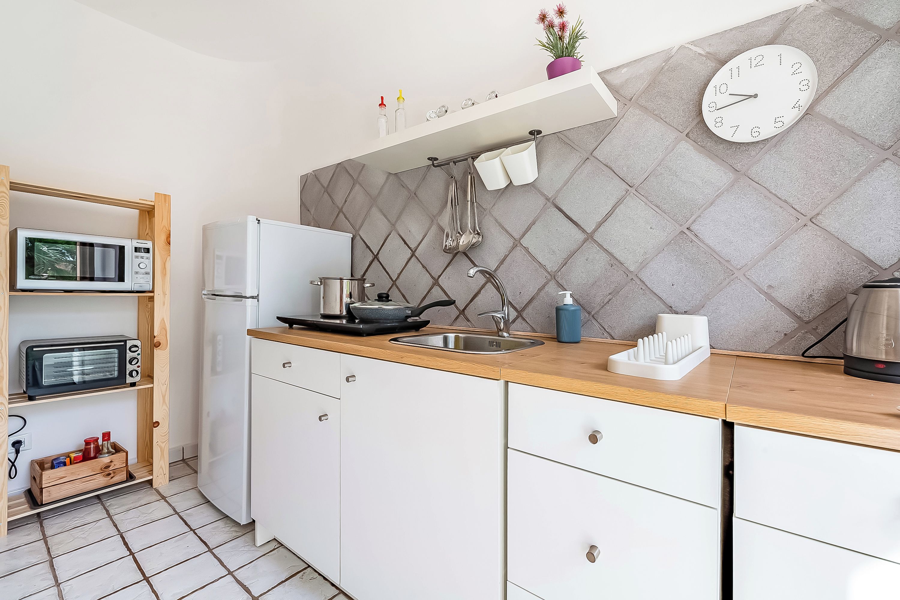 A modern kitchen with a microwave, toaster, and refrigerator, wooden cabinets, a sink, and a clock on the wall.