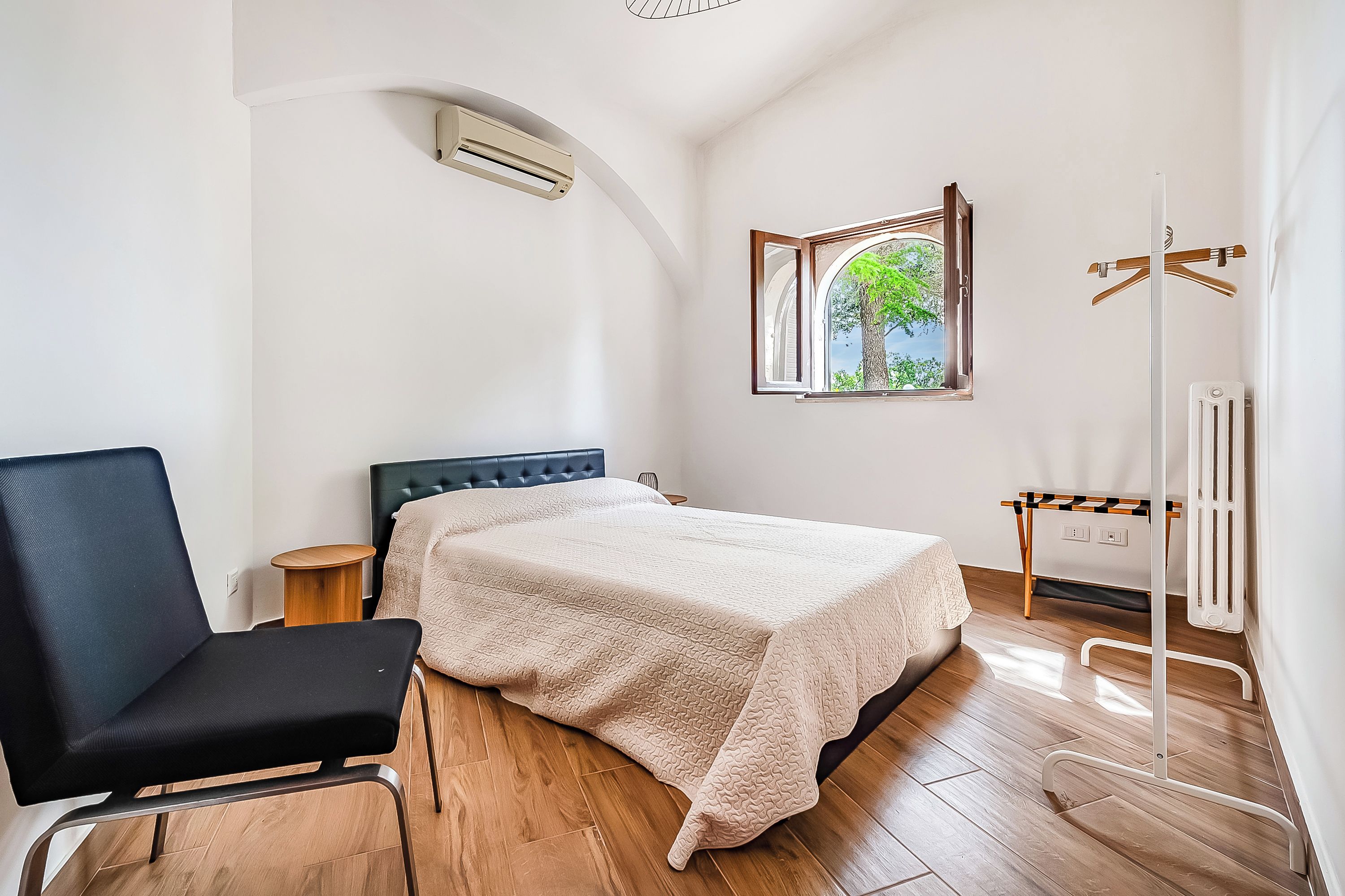 A bedroom with a bed, a chair, a radiator, and a window with a view of trees.