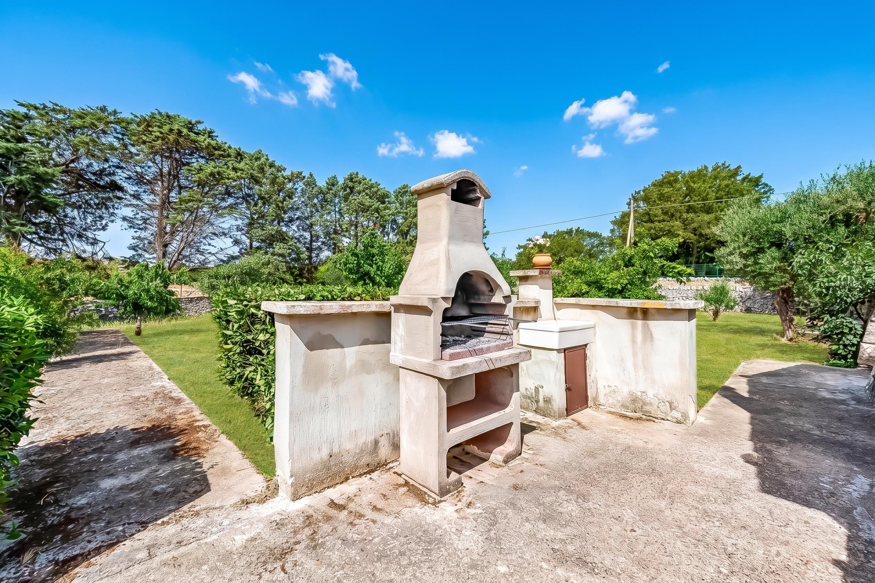 An outdoor stone oven structure with a chimney, surrounded by a well-maintained garden and a clear blue sky.