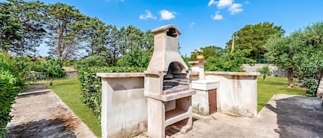 Una estructura de horno de piedra al aire libre con chimenea, rodeada de un jardín bien cuidado y un cielo azul despejado.