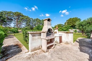 An outdoor stone oven structure with a chimney, surrounded by a well-maintained garden and a clear blue sky.
