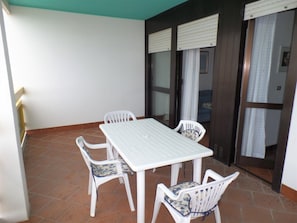 A patio with a white table and four chairs, a tiled floor, and a sliding glass door with curtains. - Beachside Apartment With Terrace (San Michele al Tagliamento)