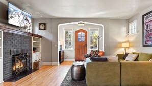 Een woonkamer met een open haard, een groene bank, een houten salontafel en een aan de muur bevestigde televisie.
