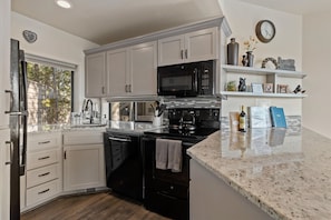 A kitchen with white cabinets, a black microwave, a stove, and a sink. There is a window with a view of trees, and a clock on the wall. - New! Deschutes River Escape with Hot Tub & Views (Bend)
