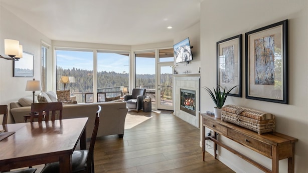 A spacious living area with a fireplace, large windows offering a forest view, and a dining area with wooden flooring. - New! Deschutes River Escape with Hot Tub & Views (Bend)