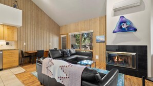 A modern living room with a fireplace, wooden walls, a black leather sofa, and a dining area. - New! Cozy Bend Escape | Hot Tub | Forest Views (Bend)