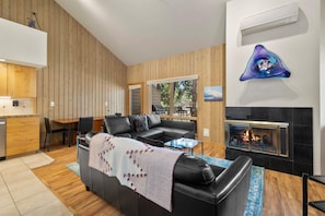 A modern living room with a fireplace, wooden paneling, a black leather sofa, and a dining area. - New! Cozy Bend Escape | Hot Tub | Forest Views (Bend)