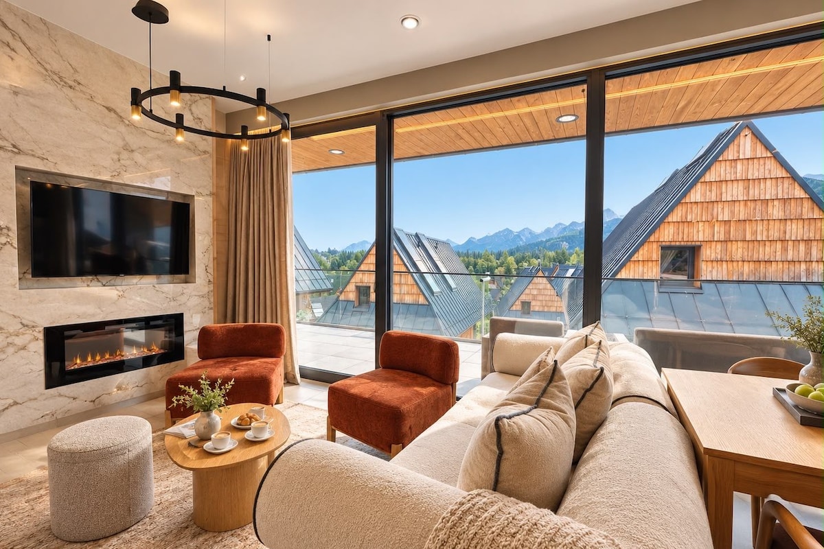 Uma sala de estar moderna com lareira, assentos confortáveis e janelas amplas com vista para casas de madeira tradicionais e montanhas.