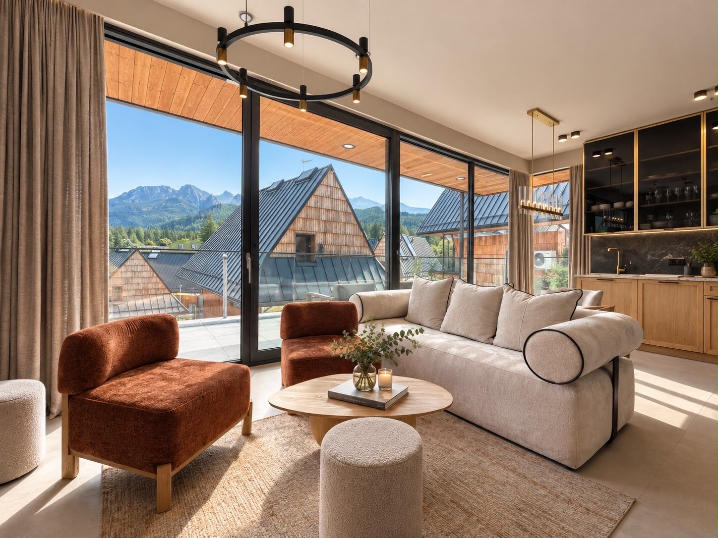 Ein modernes Wohnzimmer mit einer Couch, Sesseln und einem Couchtisch, große Fenster mit Blick auf Berge und eine Küchenzeile mit Holzschränken.
