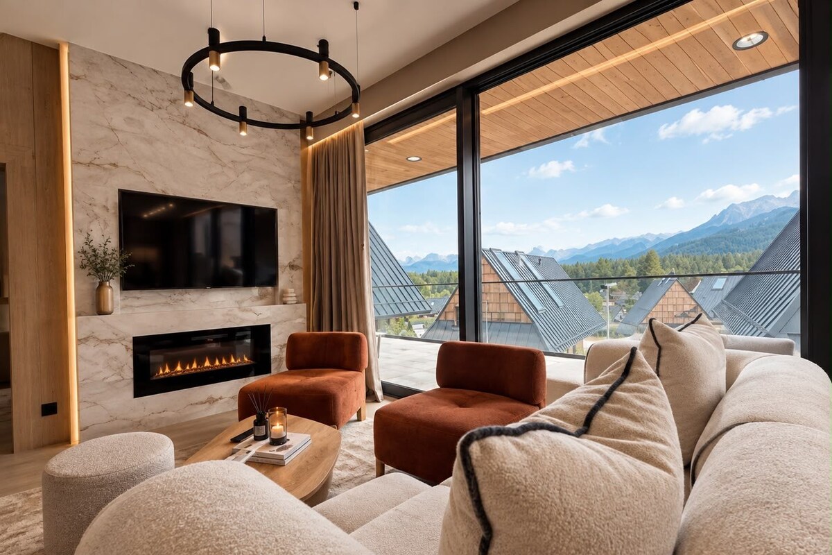 A modern living room with a fireplace, comfortable seating, a large TV, and a view of mountains.