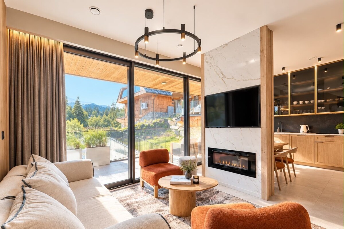 Un salon moderne comprenant un canapé, un fauteuil et une cheminée, de grandes fenêtres offrant une vue sur la montagne, ainsi qu’un coin cuisine avec des placards et un téléviseur.