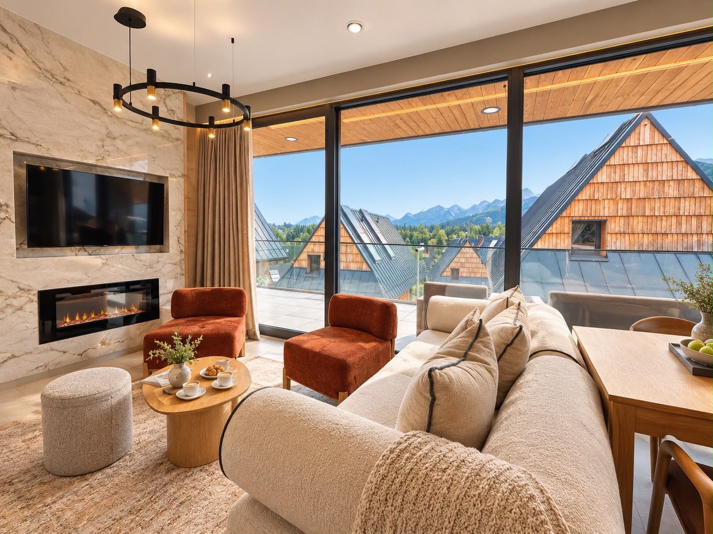A modern living room with a fireplace, comfortable seating, and large windows offering a view of traditional wooden houses and mountains.