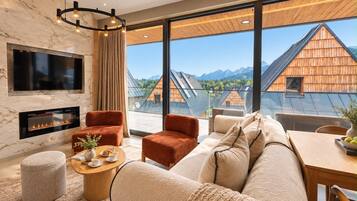 A modern living room with a fireplace, comfortable seating, and large windows offering a view of traditional wooden houses and mountains.