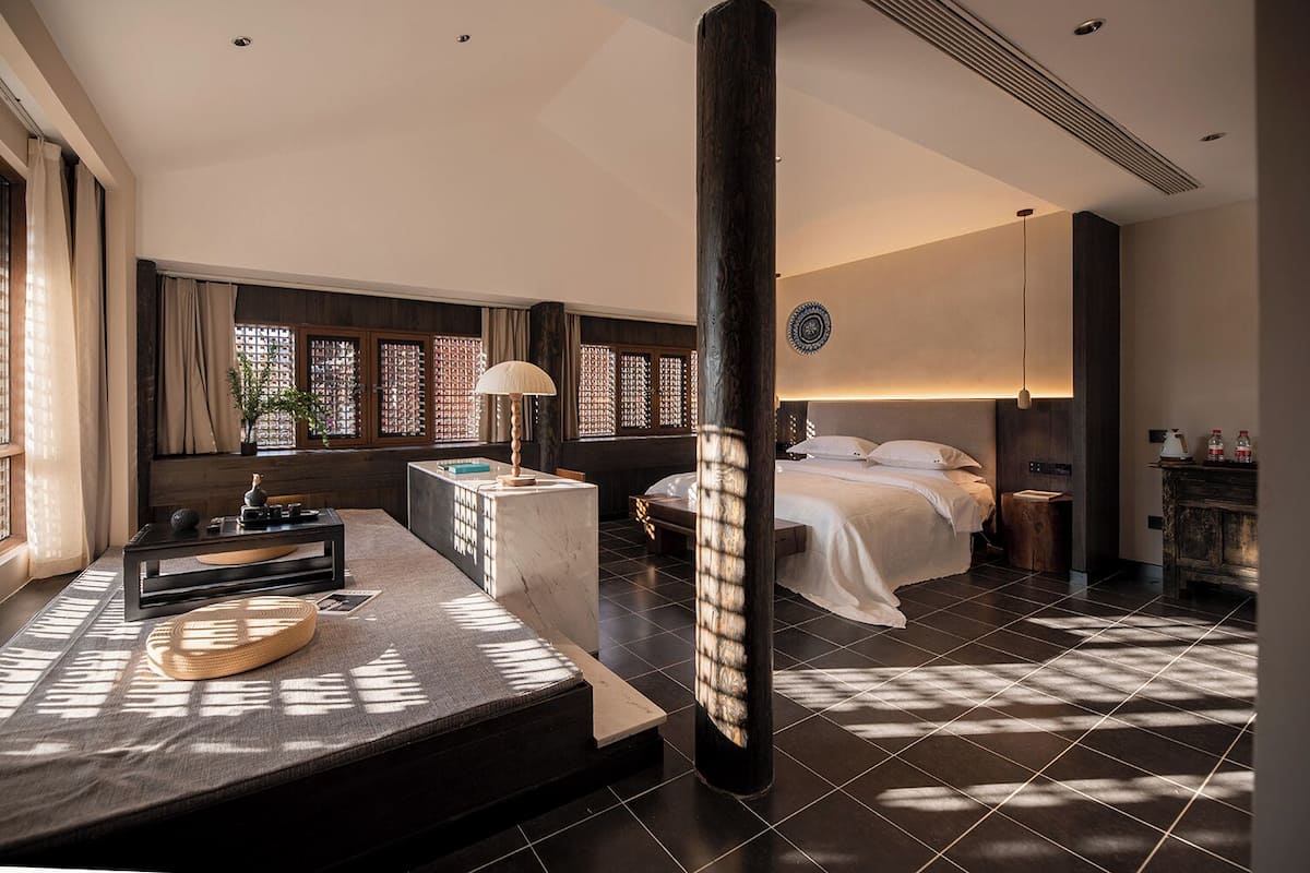 Ein modernes Hotelzimmer mit Bett, Nachttisch, Lampe und Blick durch ein mit Vorhängen versehenes Fenster.