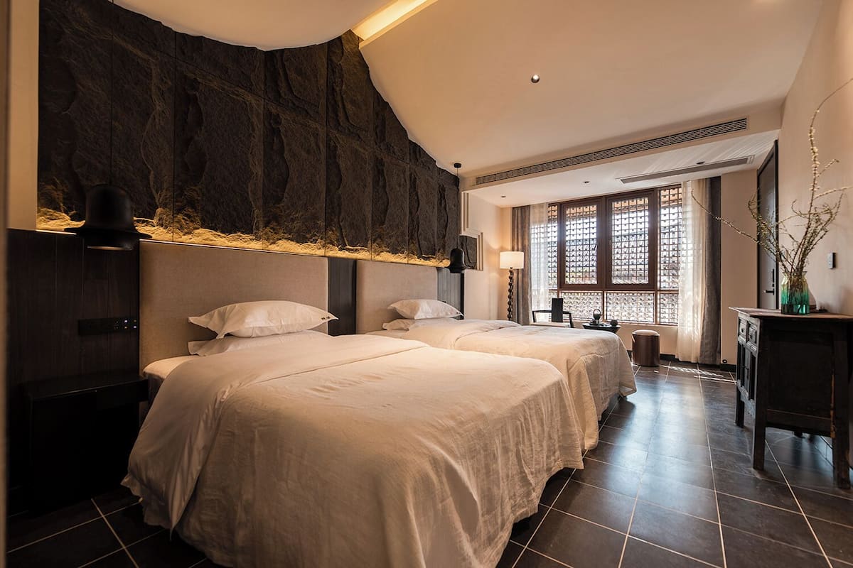Ein Hotelzimmer mit zwei Betten, einem großen Fenster mit Blick, einer dunklen Wand mit Struktur und einem kleinen Tisch mit einer Vase.