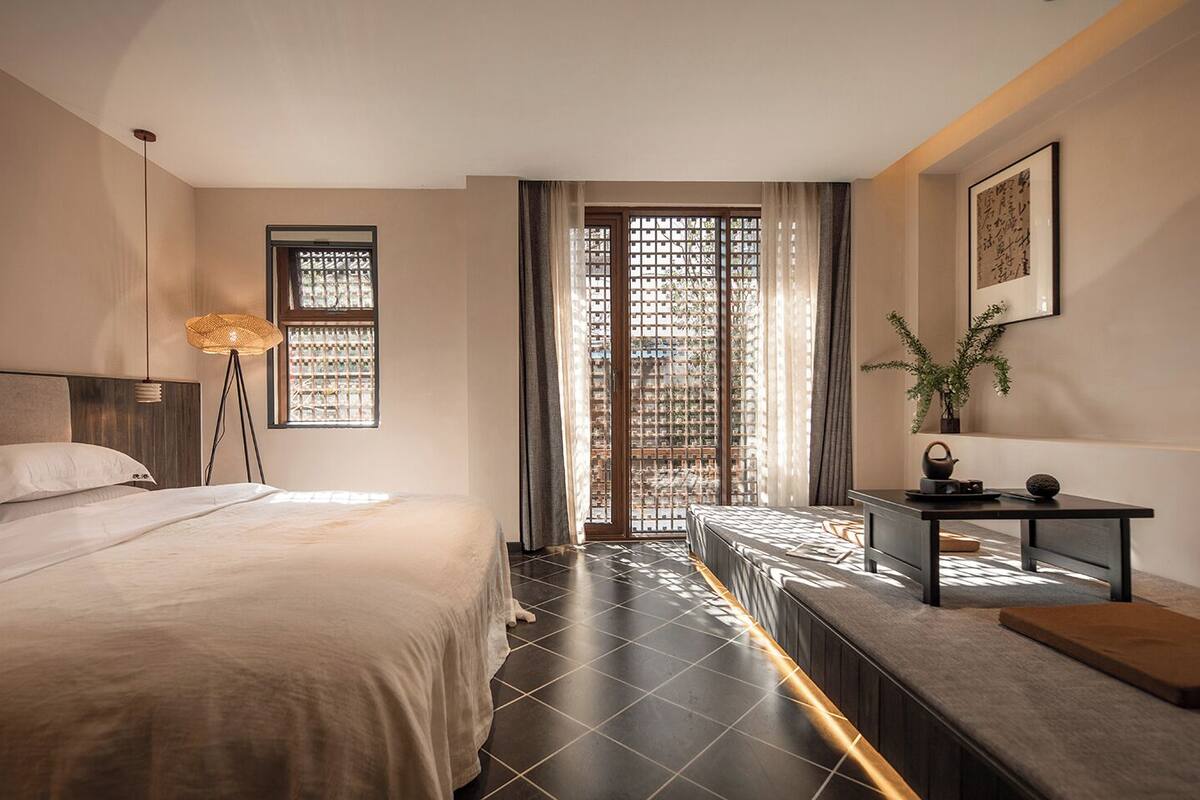 Ein modernes Hotelzimmer mit Bett, Nachttisch, Lampe und Blick durch ein Fenster mit Vorhang.