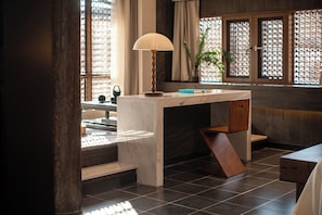 Een kamer met een houten bureau, een lamp en uitzicht door een raam met een openslaande railing.