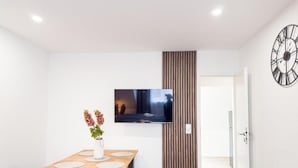 Una sala moderna con un televisor de pantalla plana montado en la pared, una mesa de comedor de madera con bancos y un jarrón decorativo con flores.
