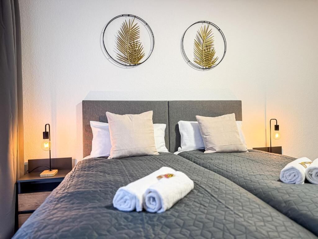 A double bed with a grey quilted cover, two white pillows, and rolled towels on the bed. There are two bedside tables with lamps, and two wall-mounted circular decorations.
