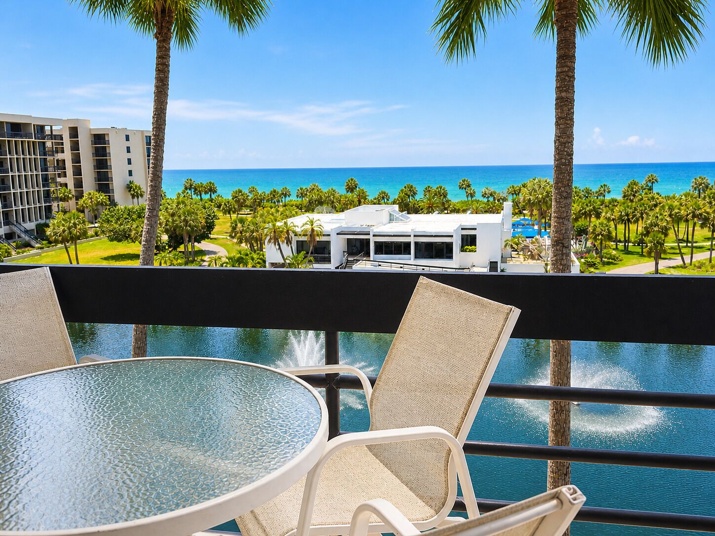 Een balkon met een glazen tafel en rieten stoelen, met uitzicht op een zwembad en een panorama van de oceaan en resortgebouwen.