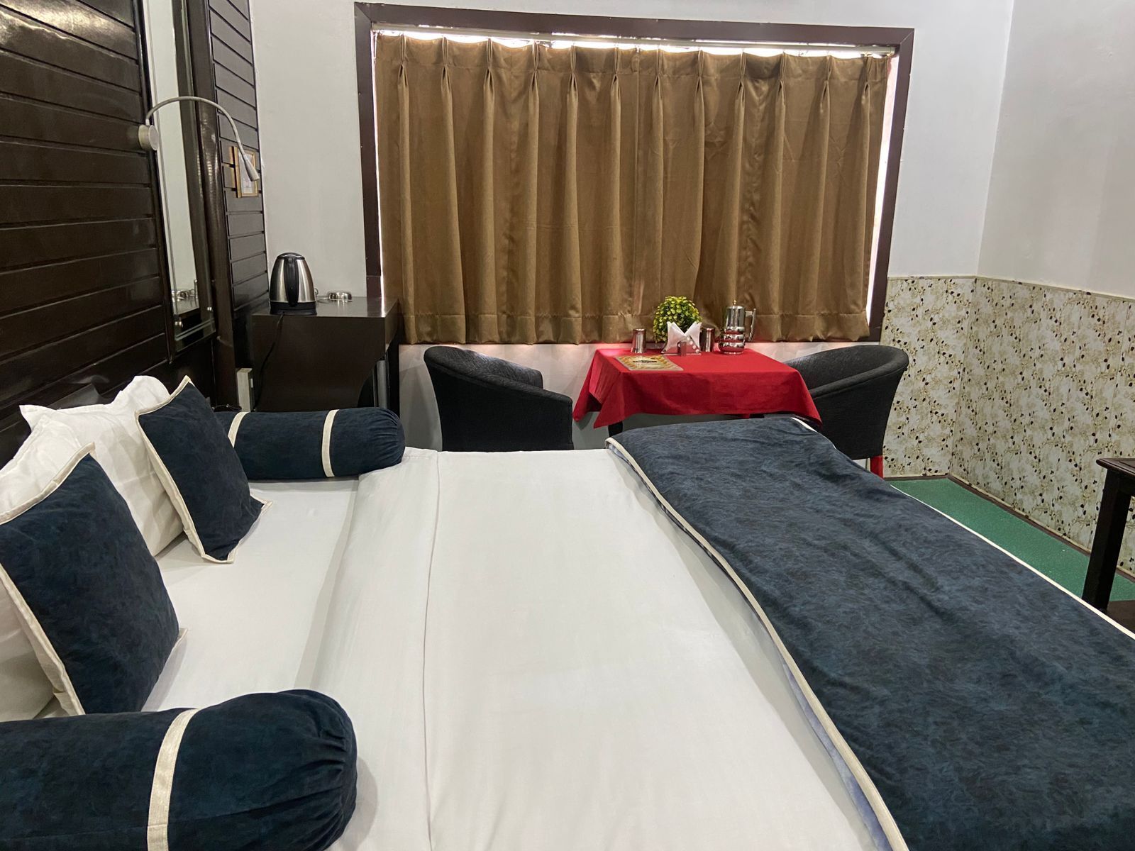 Une chambre d’hôtel avec un lit, deux tables de chevet, un bureau avec une chaise, une table rouge avec un vase et une fenêtre avec des rideaux.