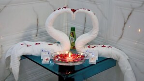 A table with a heart-shaped arrangement of white towels, a bottle of hair care product, and a bowl of rose petals. - Le-Leisure Hotel (Prayagraj)