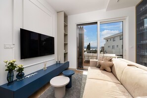 A modern living room with a flat-screen TV, a blue console table, a beige sofa, and a glass door leading to a balcony with a city view. - Deluxe 2 bedroom apartment Azzura with swimming pool and sea view (Malinska)