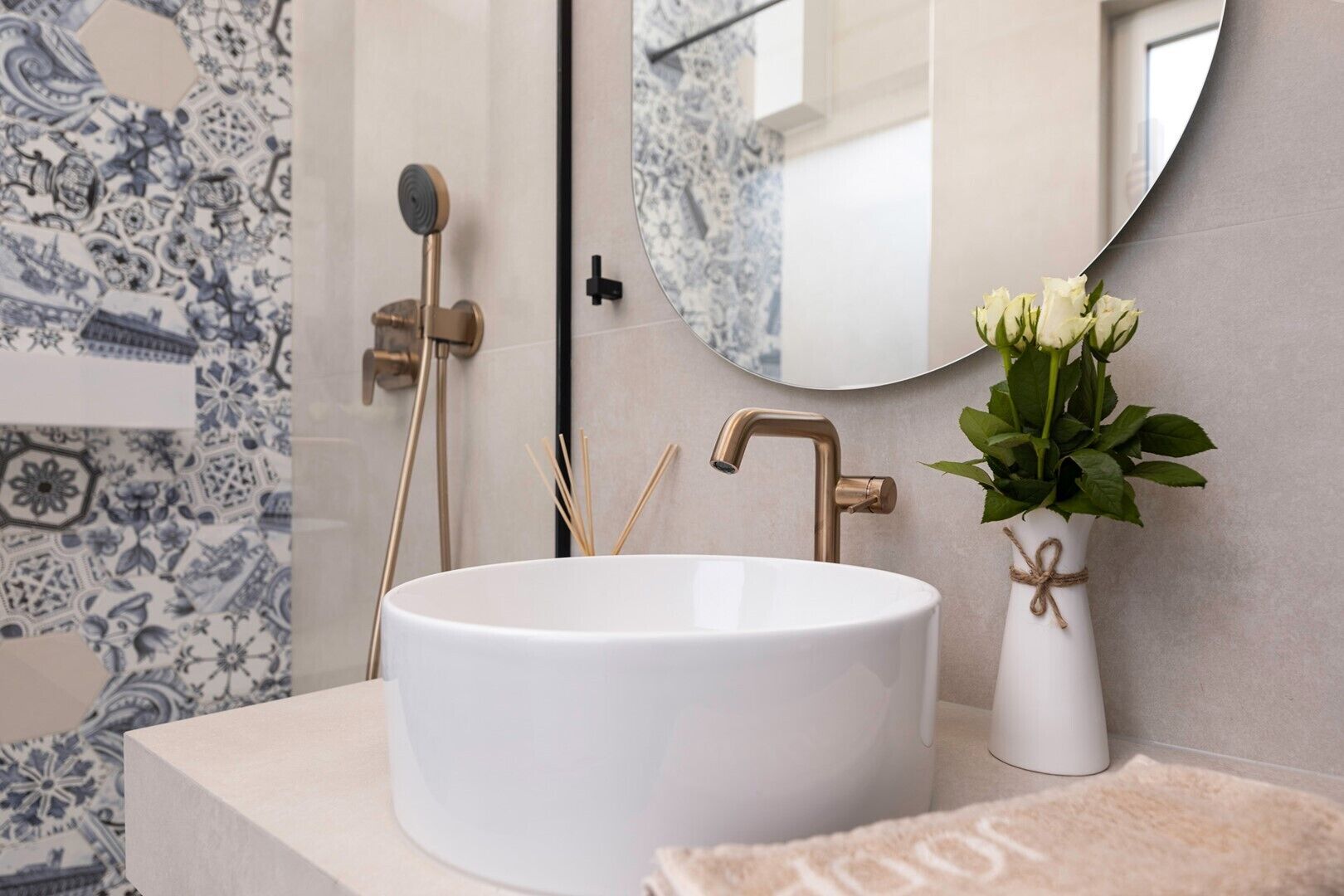 Un baño con lavamanos blanco, grifo dorado, espejo y un florero con flores blancas.
