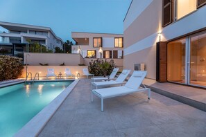 A modern outdoor pool area with lounge chairs, a clear pool, and a building with large windows and a staircase. - Deluxe Olive Apartment with three bedrooms, swimming pool, and sea view – ground floor (Malinska)