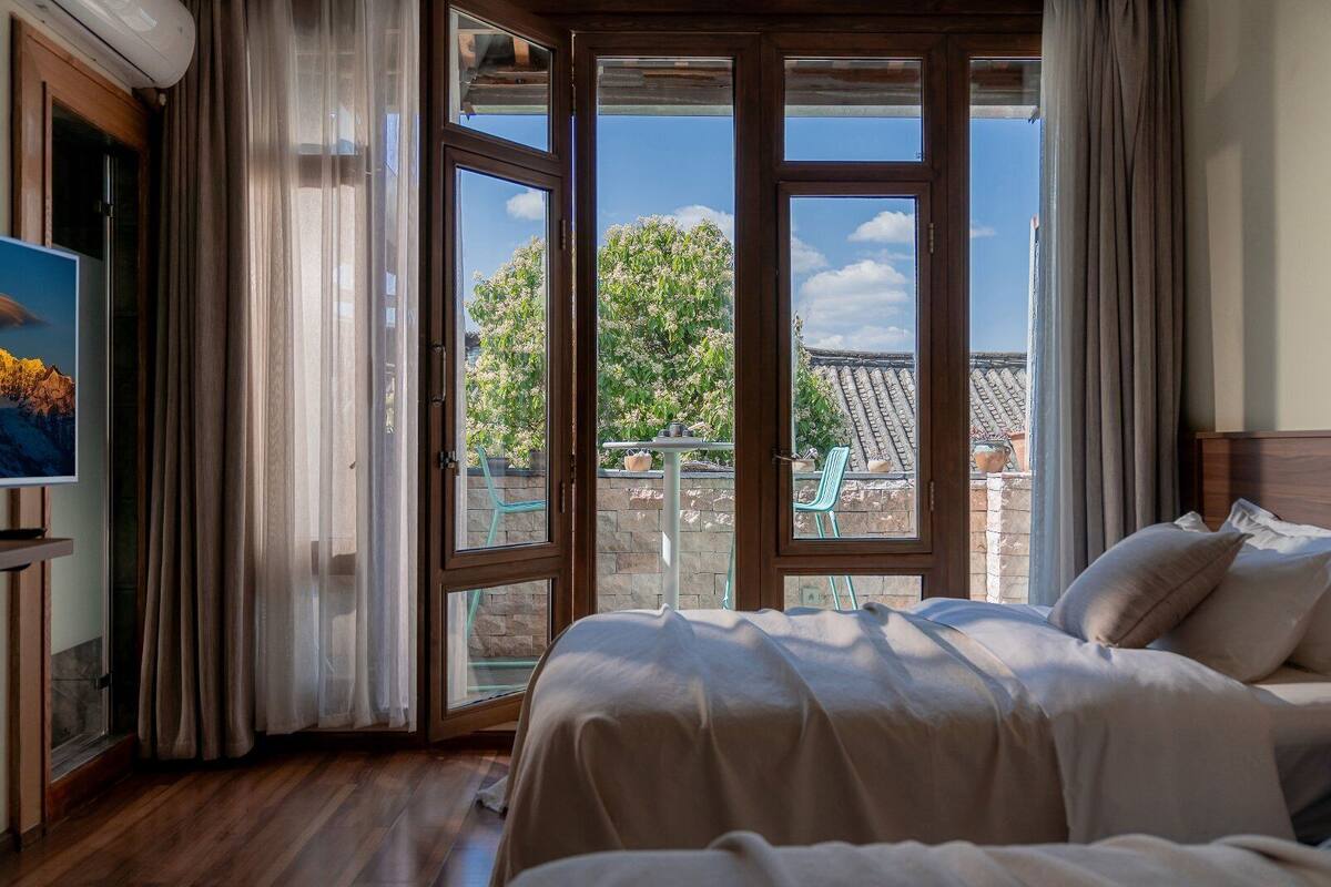 A bedroom with a bed, a TV, and a balcony with a view of a building and trees.