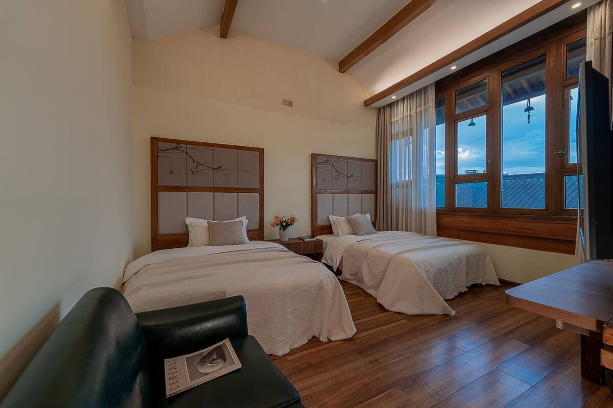 A hotel room with two beds, wooden flooring, a small table, and a window with a view.