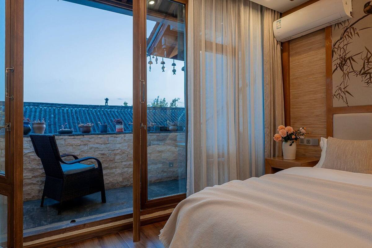 A bedroom with a bed, a chair, a nightstand with a vase, and a view of a tiled roof through a glass door.