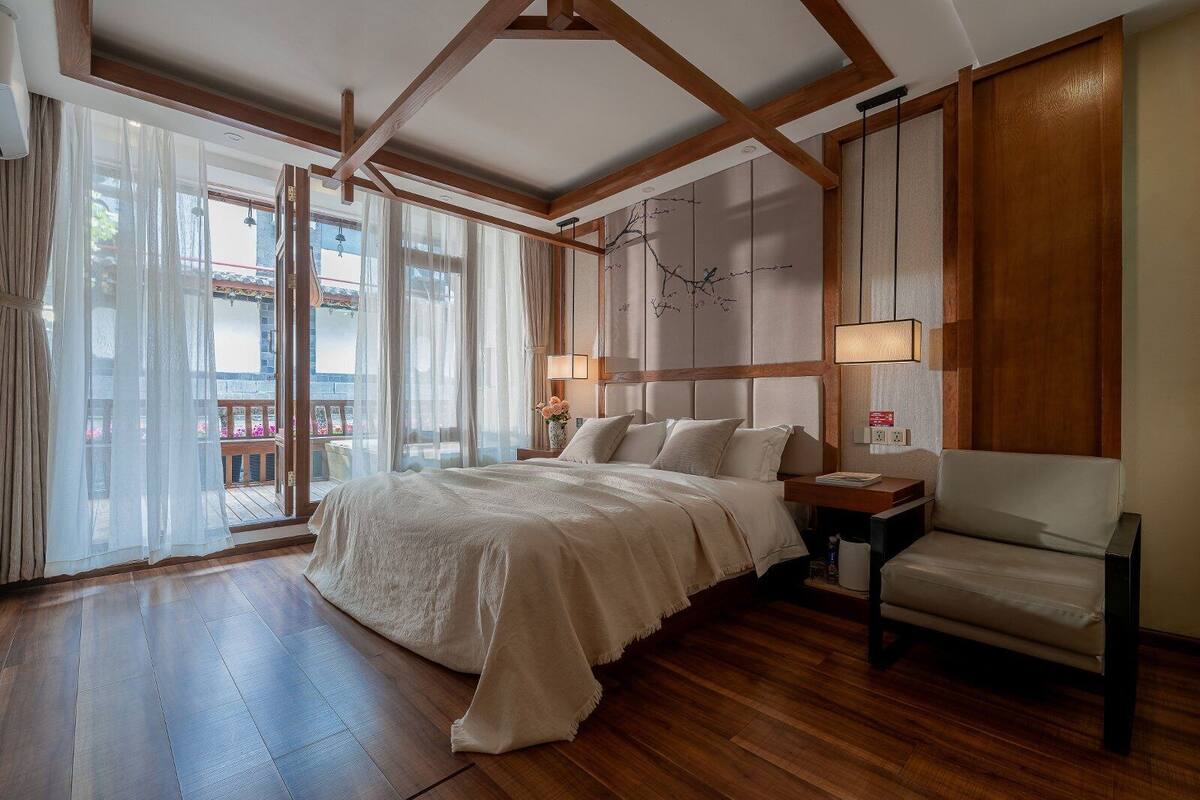 A bedroom with a large bed, a bedside table, a chair, wooden beams, a view through a window, and a wall-mounted artwork.