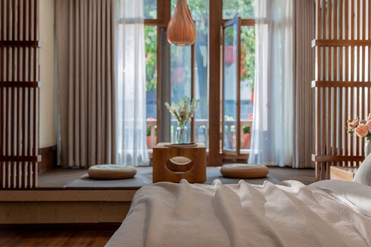 A bedroom with a large window, a wooden bedside table, and a bed with white bedding.
