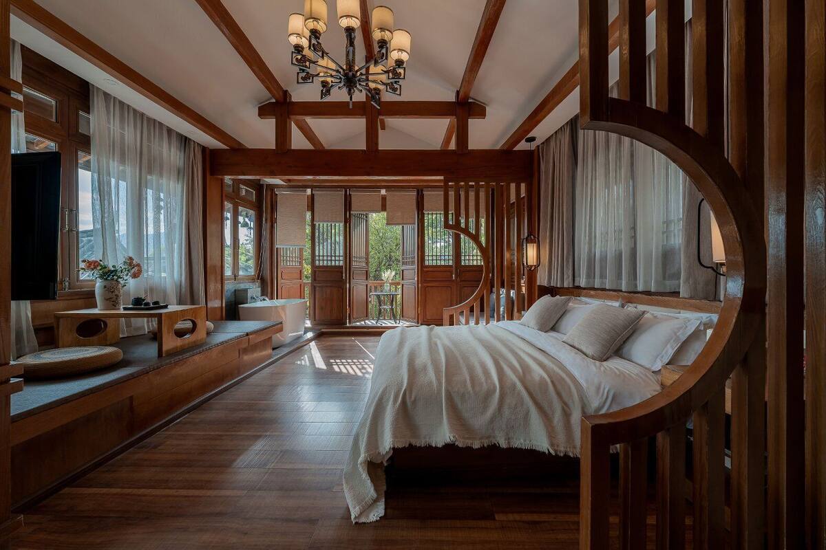 A bedroom with a large bed, wooden beams, a chandelier, and a view through a wooden-framed window.