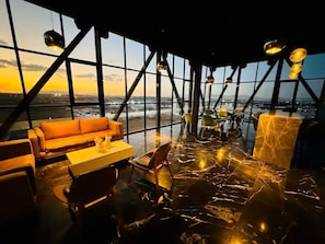 Ein hoher Innenraum mit Blick auf die Stadt, eine Couch, ein Esstisch, Stühle und hängende Pendelleuchten.