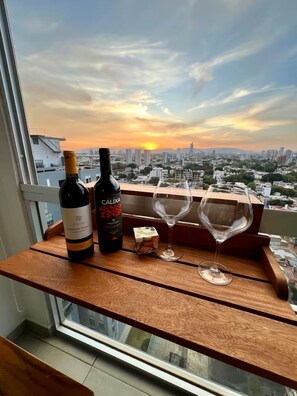 Ein Tisch mit einer Monte Xanic Weinflasche, einer Calixa Flasche und zwei Weingläsern auf einem Balkon mit Blick auf eine Stadtlandschaft bei Sonnenuntergang.
