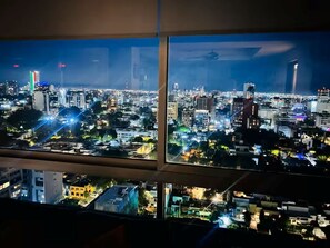 Blick auf eine Skyline aus einem Fenster, mit beleuchteten Gebäuden und einem Hubschrauber am Himmel.