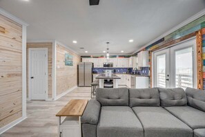 A modern kitchen with white cabinets, a central island, and a dining area with a grey sofa and wooden table. - CL3 Shared Pool, Charcoal Grill, Old Town Port A (Port Aransas)