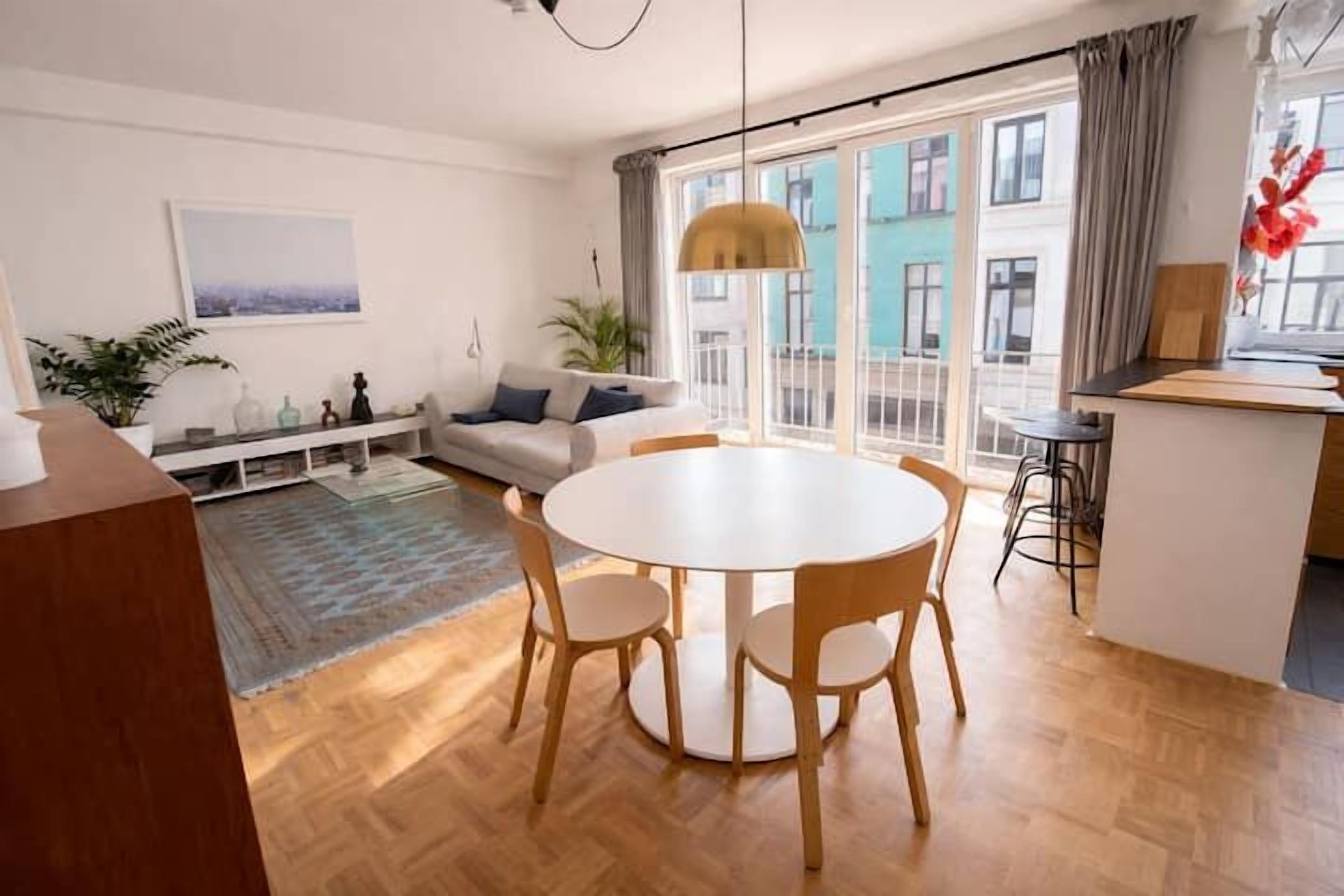 A modern living room with a round dining table, a sofa, a small kitchen area, and large windows with a city view.