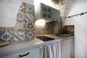 A kitchen with a stone wall, a built-in stove, a hanging pot rack, and a tiled backsplash. - Cozy apartment on two levels that accommodate up to 3 people and is equipped with all the main comforts for a pleasant stay. The location is excellent, in the center, with all the essential services at hand and a short distance from the points of interes (Martina Franca)